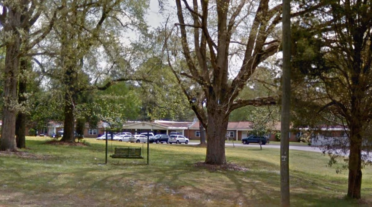 Outdoor view of Magnolia Gardens of Warrenton showing a grassy area with large trees, a green metal swing bench, and a parking lot with several cars in front of a single-story brick building.