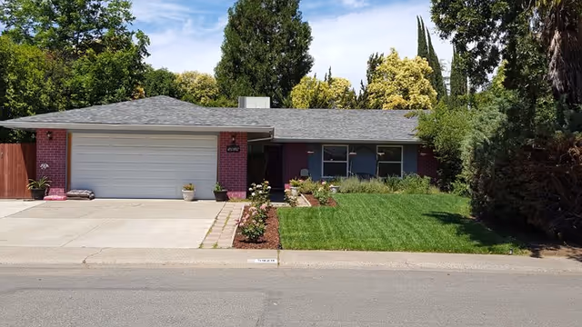 Front exterior view of a single-story brick house with a two-car garage, a well-maintained lawn, flower beds, and trees in the background under a partly cloudy sky.