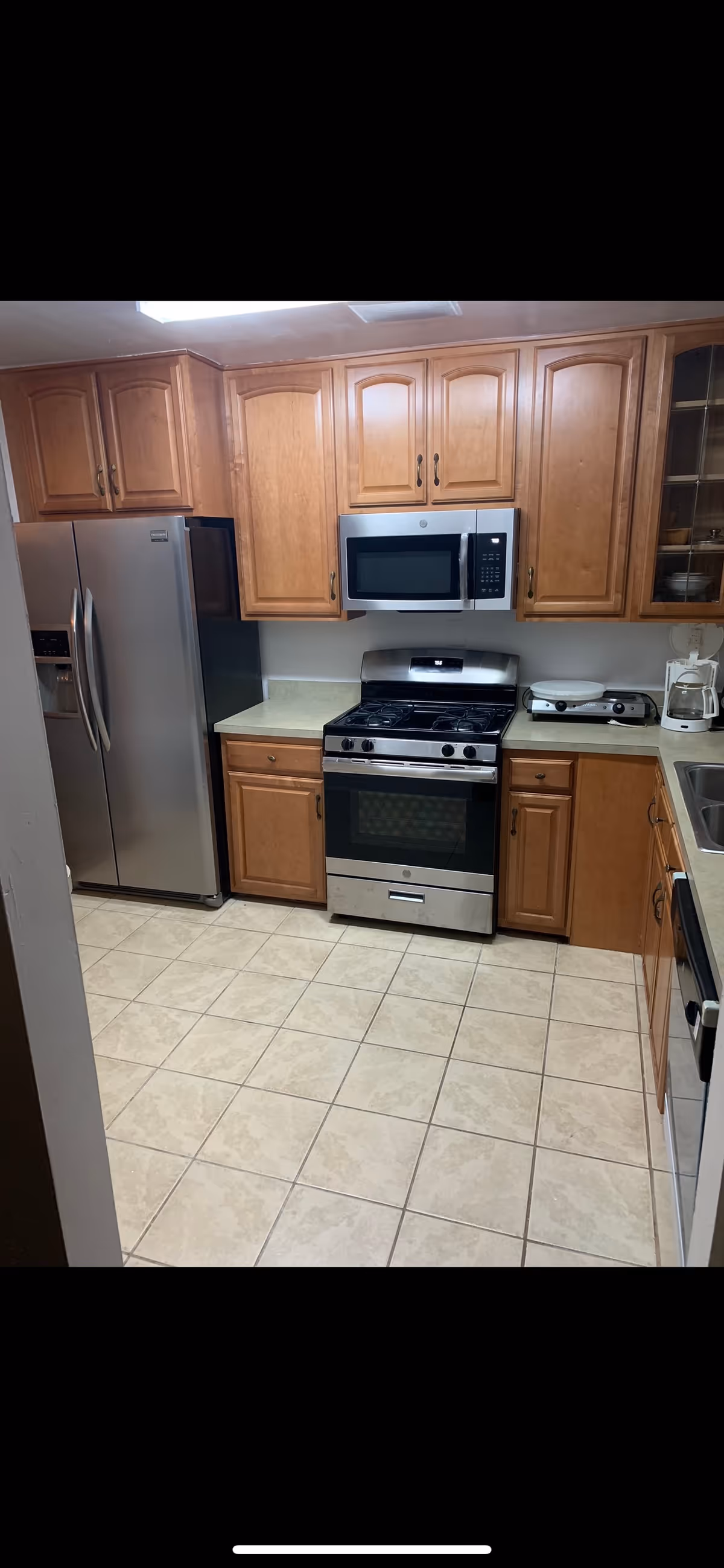 A kitchen with wooden cabinets, a stainless steel refrigerator, a stainless steel gas stove with oven, a microwave above the stove, a countertop with a coffee maker and a double burner hot plate, and a tiled floor.