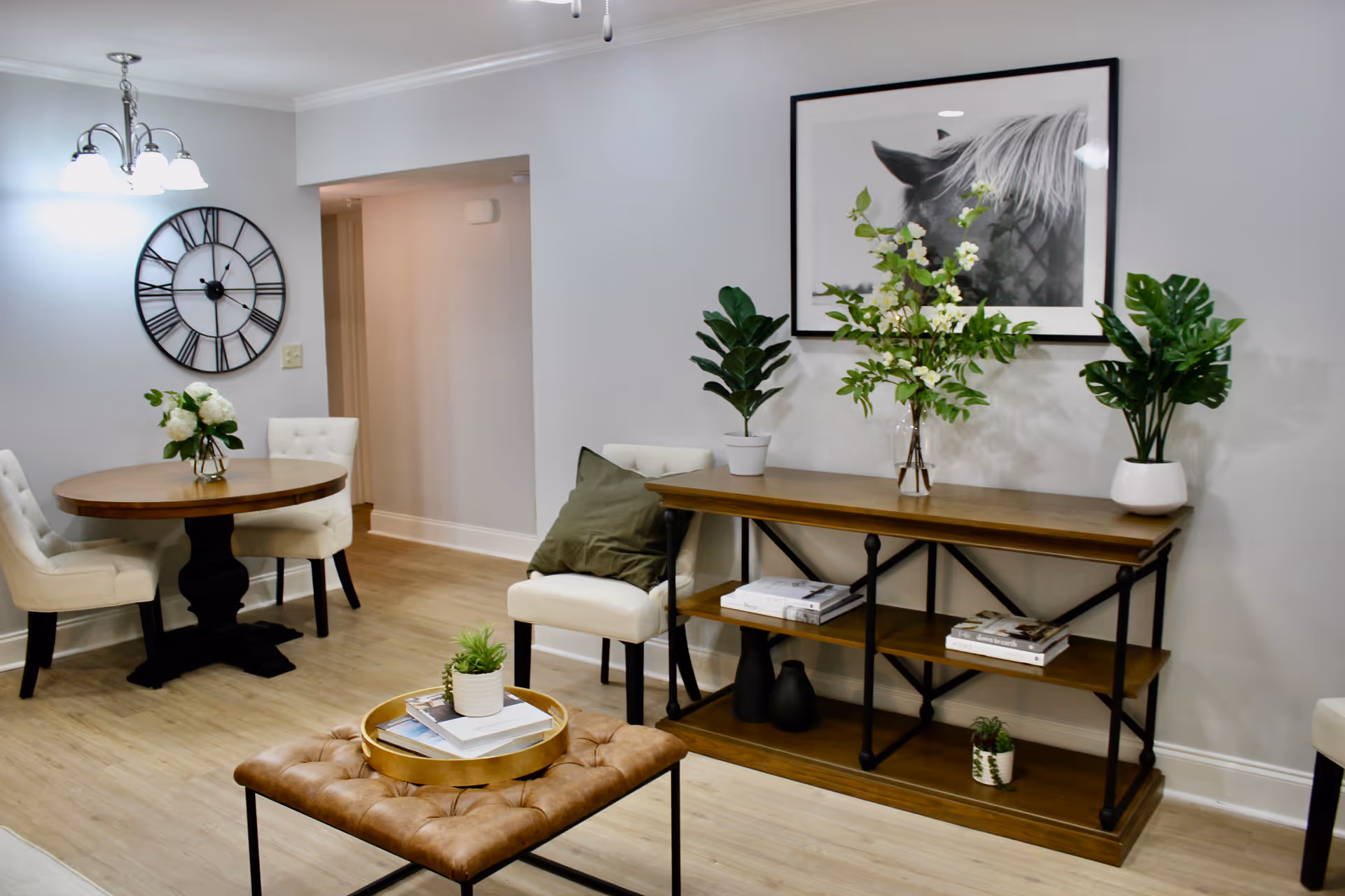 A cozy interior space featuring a round wooden dining table with white cushioned chairs, a large wall clock, a wooden console table with potted plants and books, a framed black and white horse photograph on the wall, and a tufted ottoman with a tray and small plant on it.
