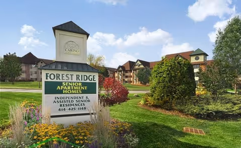 Exterior view of Forest Ridge Senior Apartment Homes showing a large sign with the facility name and contact information, surrounded by well-maintained landscaping with flowers, bushes, and trees, and buildings in the background under a partly cloudy sky.