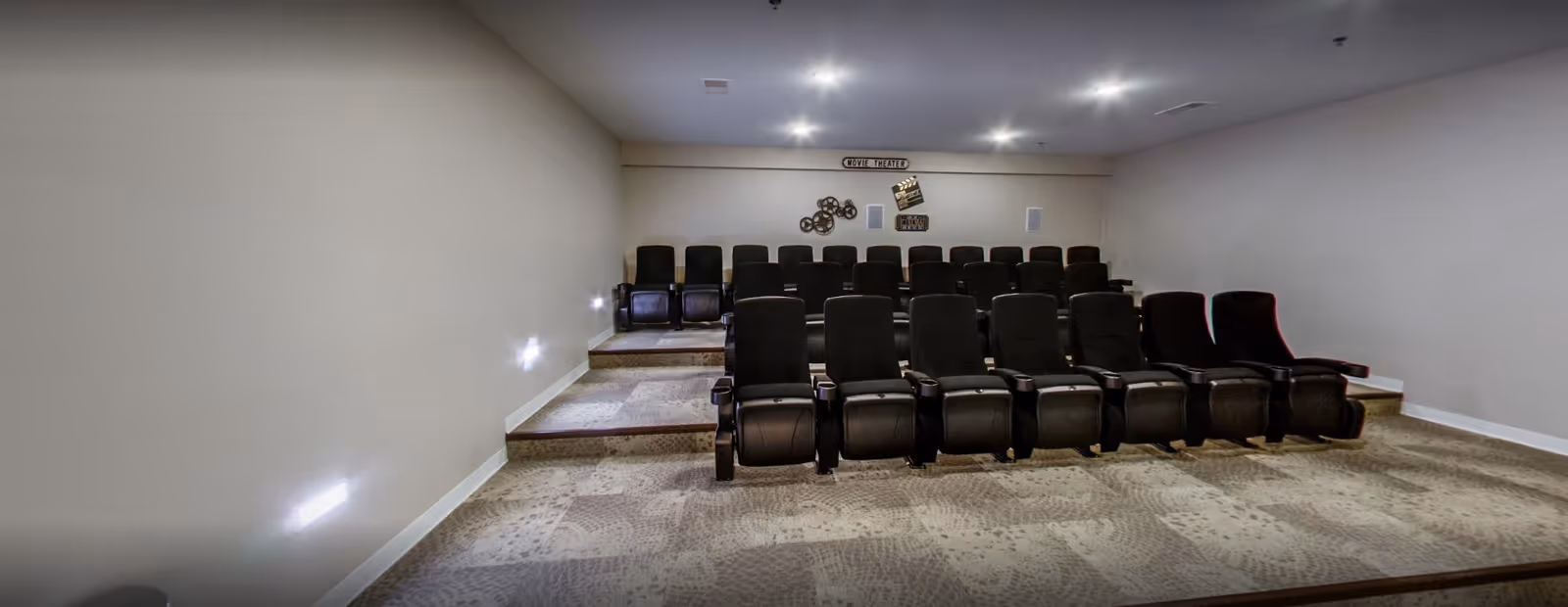 A small movie theater room with three rows of black cushioned seats arranged on carpeted steps. The walls are plain and light-colored, with a few movie-themed decorations on the back wall, including a sign that says 'MOVIE THEATER' and film reel and clapperboard decor. The room is softly lit with recessed ceiling lights and wall lights along the steps.