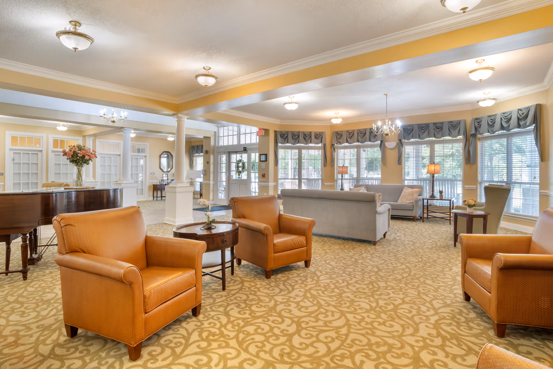 A spacious and well-lit senior living facility common area with patterned carpet, several comfortable armchairs and sofas, a grand piano with a vase of flowers on top, large windows with blue valances allowing natural light, and multiple ceiling light fixtures.
