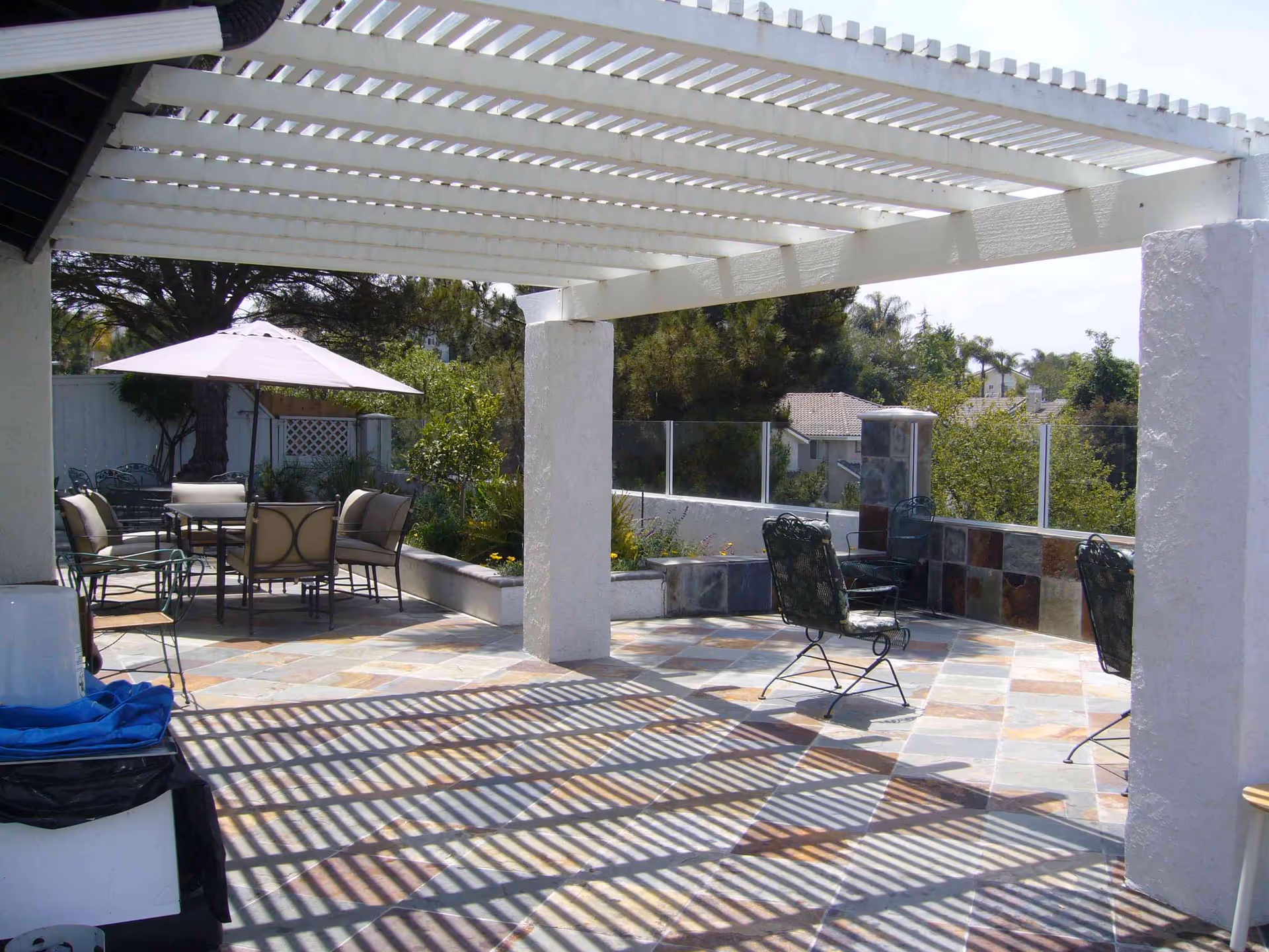 Outdoor patio area with a white pergola casting shadows on the tiled floor. There is a seating area with cushioned chairs and a table under a large umbrella, and additional metal chairs arranged around the patio. Trees and greenery are visible in the background.
