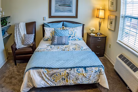 A neatly made bedroom with a twin bed dressed in patterned bedding, an armchair, nightstand with lamp, and a window with blinds.