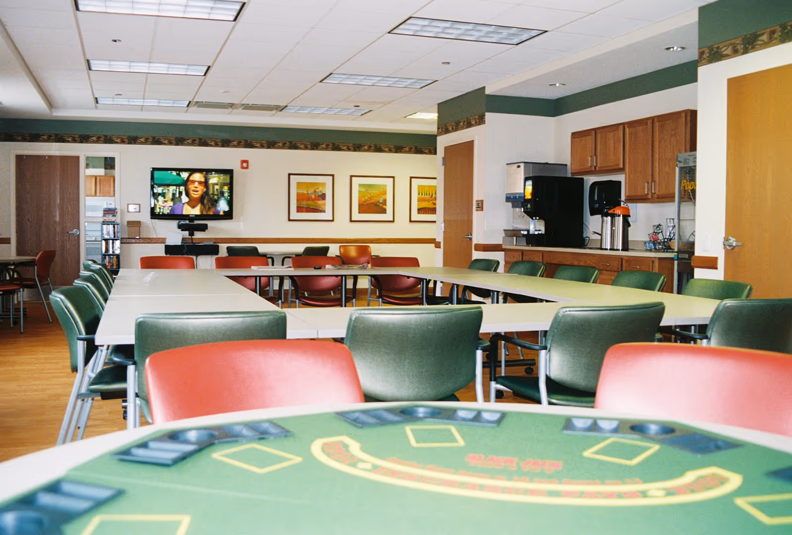 A spacious room with multiple tables and chairs arranged for group activities or meetings. The chairs are a mix of green and red colors. There is a television mounted on the far wall displaying a person speaking. The room has wooden flooring, framed artwork on the walls, and a counter area with a coffee machine and other supplies.