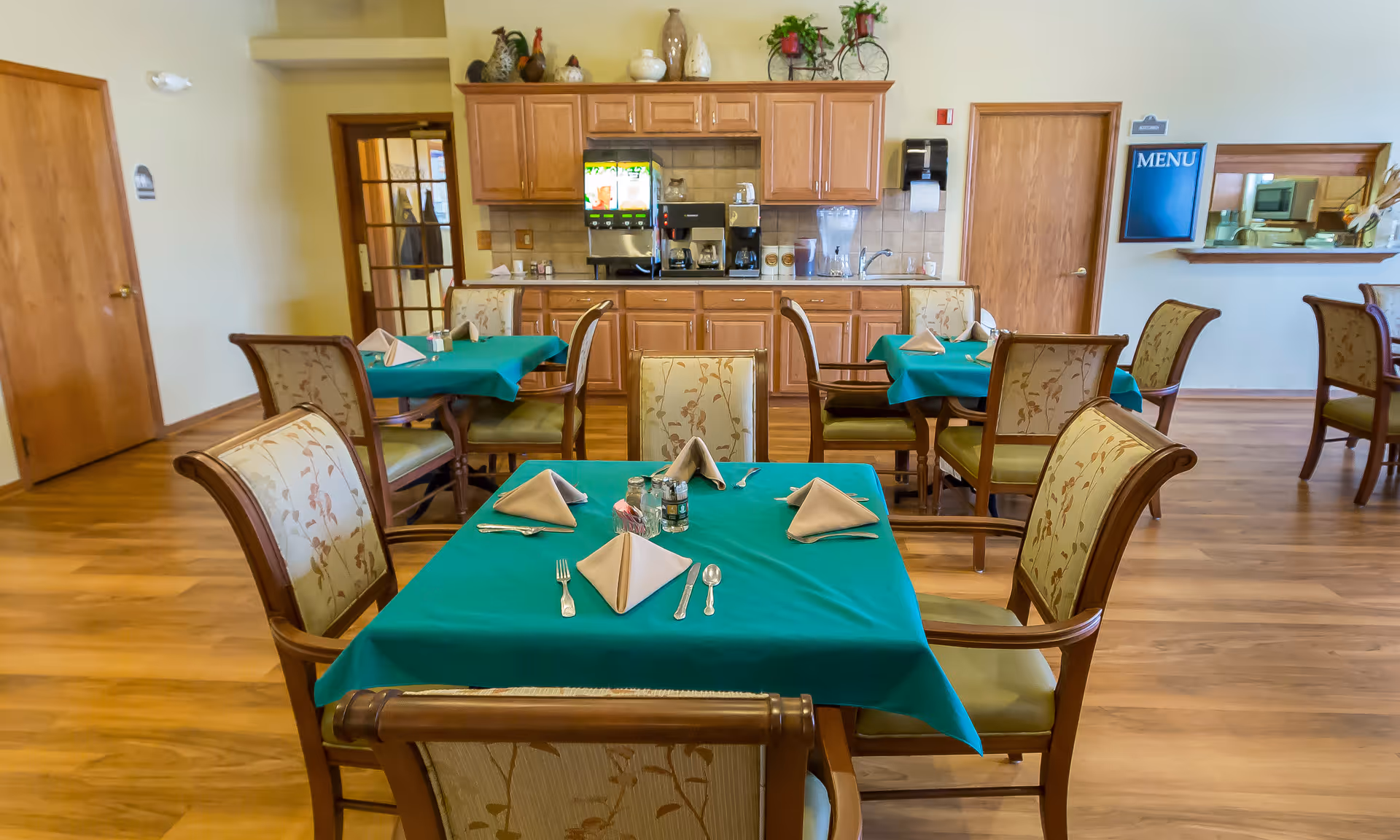Dining area in a senior living facility with tables set with green tablecloths and folded napkins and a kitchenette/counter in the background.