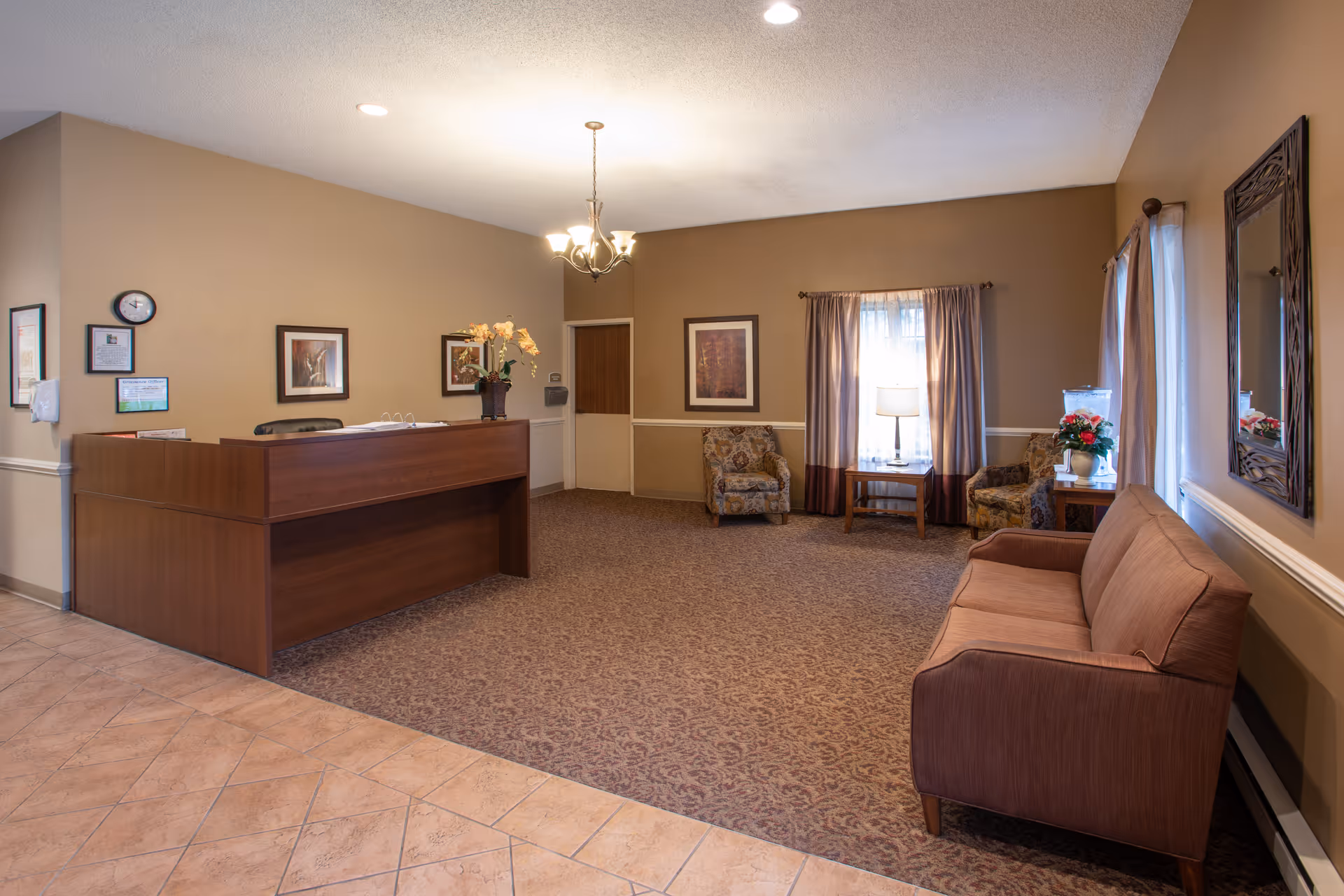 Reception area of New Castle Health & Rehabilitation Center featuring a wooden front desk with a vase of flowers, a wall clock, framed pictures, a brown sofa, two patterned armchairs, a side table with a lamp, and windows with curtains.