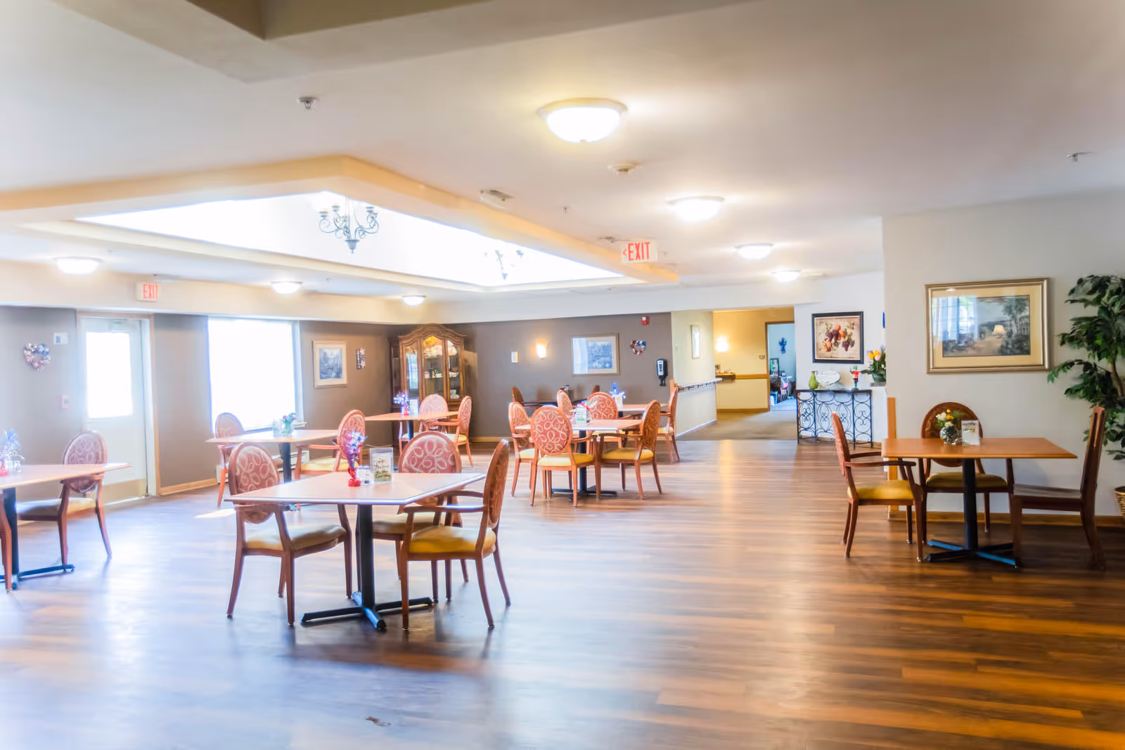 A bright and spacious dining area in an assisted living facility with several wooden tables and chairs arranged neatly. The room has large windows allowing natural light, wooden flooring, framed artwork on the walls, and decorative plants. Ceiling lights and an exit sign are visible.