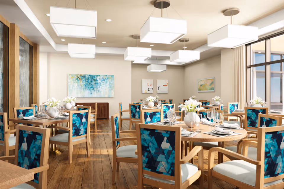 A bright and modern dining room with wooden floors and multiple round and square tables set with white plates, glassware, and floral centerpieces. The chairs have wooden frames with blue patterned upholstery on the backrests. Large windows on the right side let in natural light, and contemporary rectangular pendant lights hang from the ceiling. Abstract artwork decorates the walls.