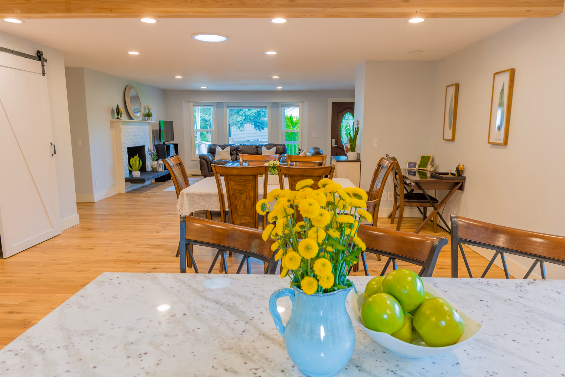 Open-plan kitchen and dining area with a marble island featuring a vase of yellow flowers and a bowl of green apples, with a dining table and living room visible in the background.