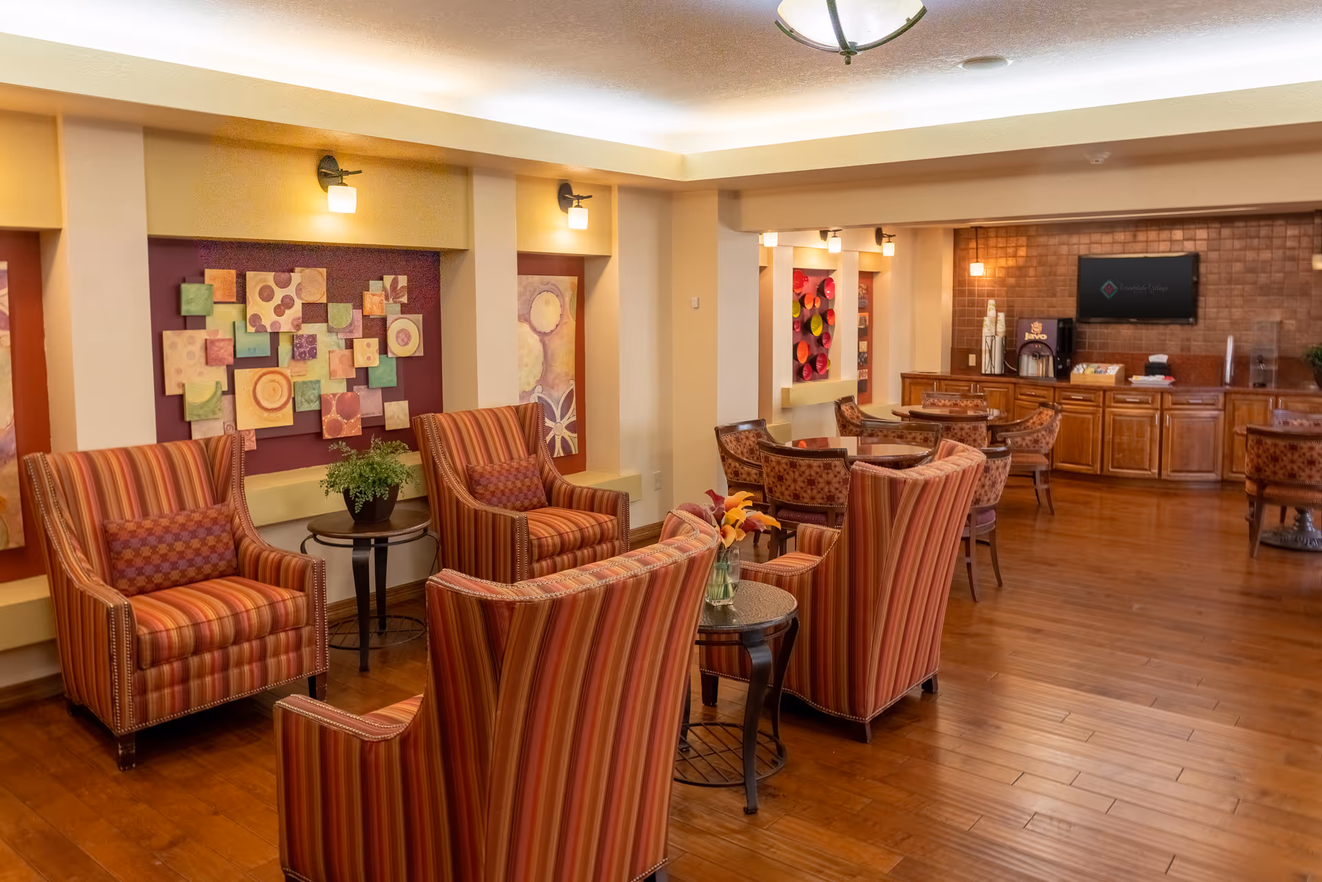 A cozy common area in a senior living facility featuring several striped upholstered armchairs arranged around small round tables with flower vases. The room has warm wooden flooring, colorful abstract wall art, soft lighting from wall sconces and ceiling fixtures, and a counter area with a coffee machine and a wall-mounted TV in the background.