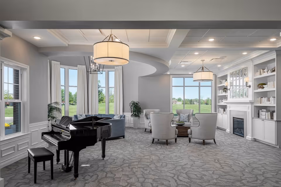 A spacious and elegant living room with large windows letting in natural light. The room features a black grand piano with a matching bench on the left side, several comfortable armchairs arranged around a wooden coffee table, and a built-in white bookshelf surrounding a fireplace on the right. The walls are painted light gray, and the carpet has a subtle floral pattern. Two large pendant lights hang from the ceiling, and there are potted plants near the windows.