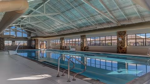Indoor swimming pool area with large windows letting in natural light, stone pillars, and a high ceiling with exposed beams and ductwork. The pool has metal handrails and steps leading into the water.