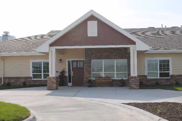 Front exterior view of a single-story building with a covered entrance supported by two columns. The building has a combination of brick and siding exterior, with three windows visible and a bench placed under the middle window. The driveway curves in front of the entrance.