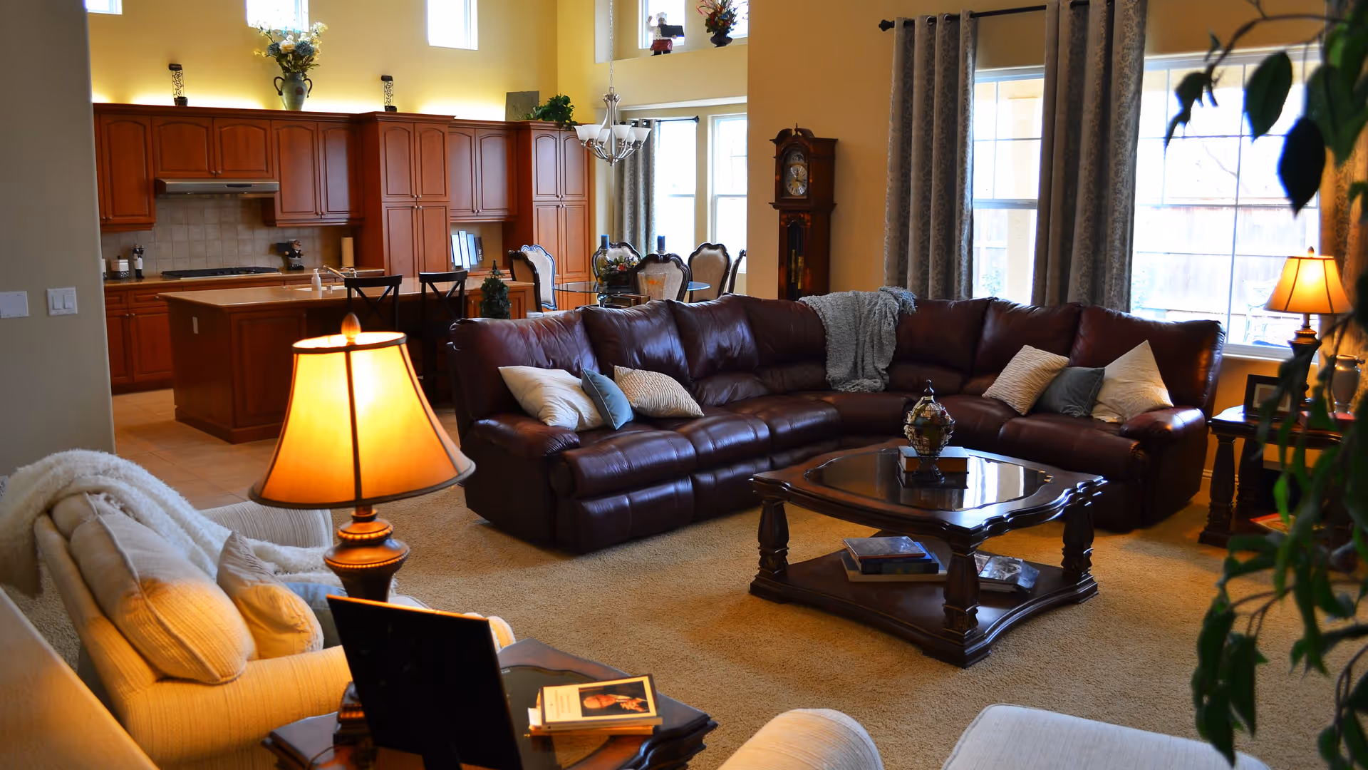 A cozy living room with a large brown leather sectional sofa adorned with multiple pillows and a throw blanket. There is a wooden coffee table with books and decorative items in front of the sofa. To the left, there is a beige armchair with cushions and a lit table lamp on a side table. The room has large windows with curtains allowing natural light to enter. In the background, an open kitchen with wooden cabinets and a dining area with chairs is visible. The walls are painted a warm beige color, and there is a grandfather clock near the windows.