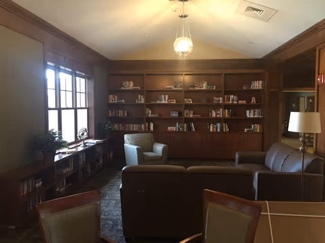 A cozy library or lounge area with wooden bookshelves filled with books along the back wall. There are two armchairs and a sofa arranged around a central space, with a floor lamp to the right. Large windows on the left side let in natural light, and there are additional bookshelves and plants beneath the windows. The ceiling has a hanging light fixture.
