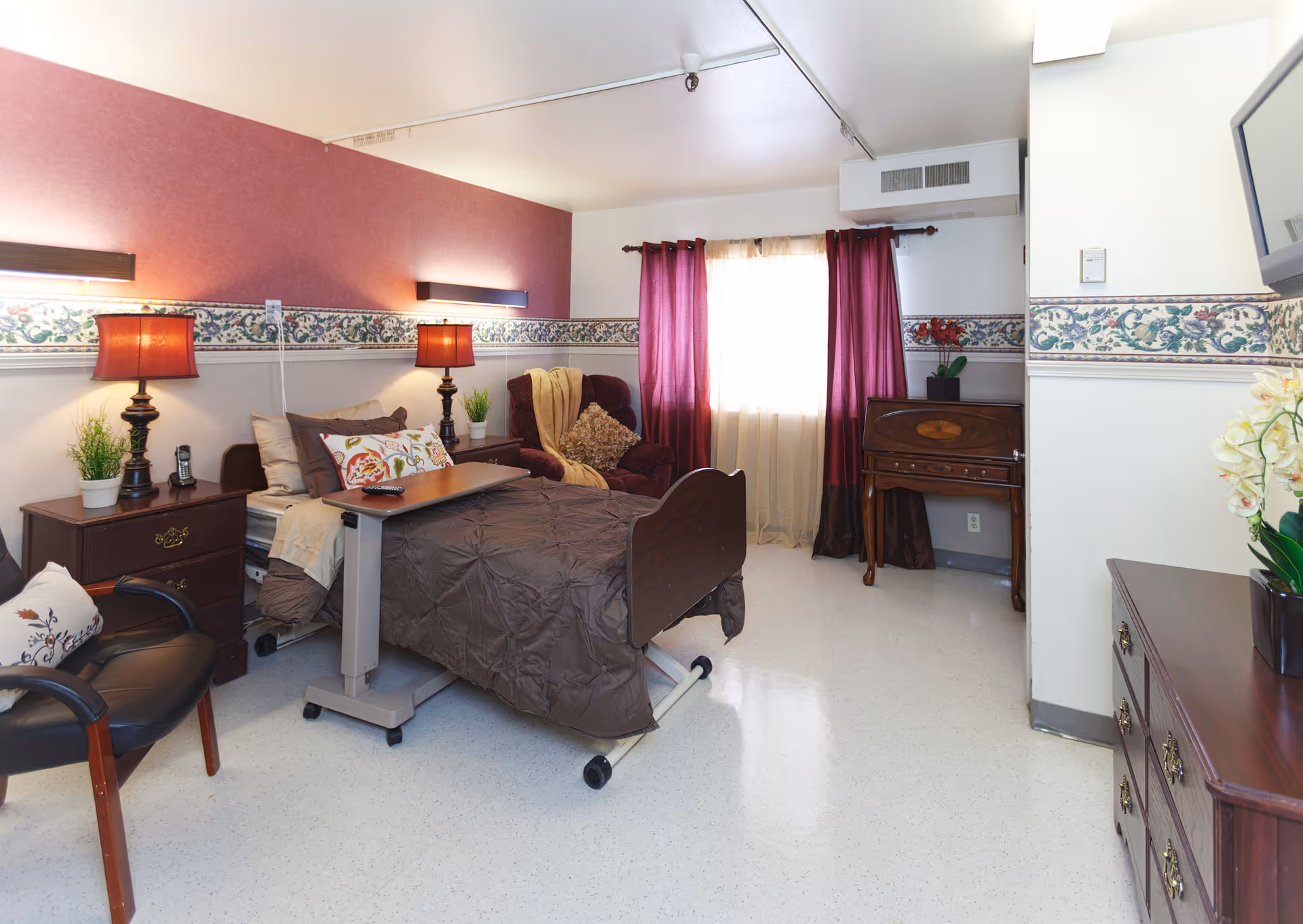 A cozy senior living bedroom with a hospital-style bed covered in a brown quilt, a small overbed table, two bedside tables with lamps and plants, a comfortable armchair with a throw blanket and pillow, a wooden desk, and a window with burgundy curtains letting in natural light.
