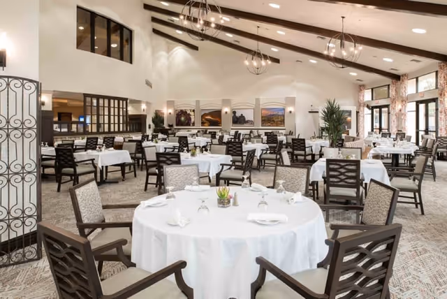 Spacious dining room with multiple round and rectangular tables covered with white tablecloths, set with plates, glasses, and napkins. The room features high vaulted ceilings with modern chandeliers, large windows allowing natural light, and framed landscape artwork on the walls.