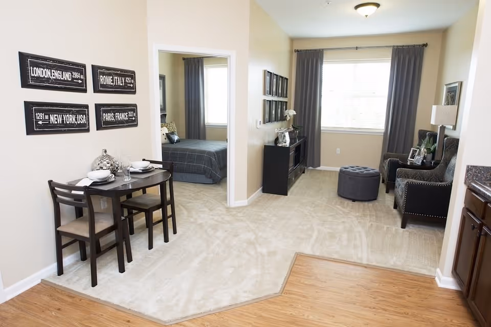 Interior view of an assisted living apartment showing a small dining area with a table set for two, a living room with two armchairs, a round ottoman, and a TV stand, and a bedroom visible through an open doorway. The walls are decorated with framed signs and pictures, and the room has beige carpet and wood flooring near the kitchen area.
