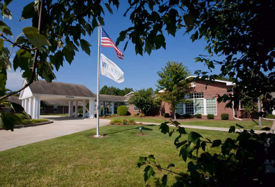 Exterior view of The Laurels of Salisbury senior living facility showing a brick building with large windows, a covered entrance driveway, two flagpoles with the American flag and a facility flag, and green lawn with trees and bushes framed by tree branches.