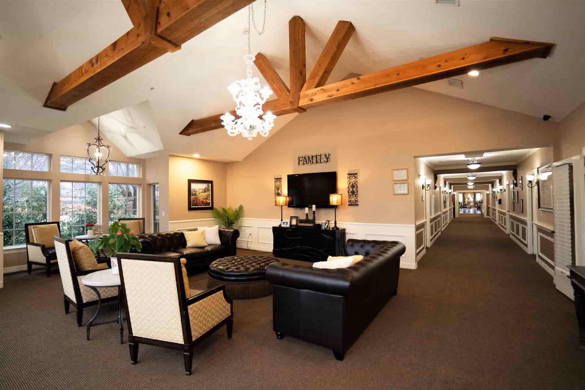 Comfortable communal living room in a senior facility with leather sofas, armchairs, a TV on the wall, exposed wooden ceiling beams and a long hallway.