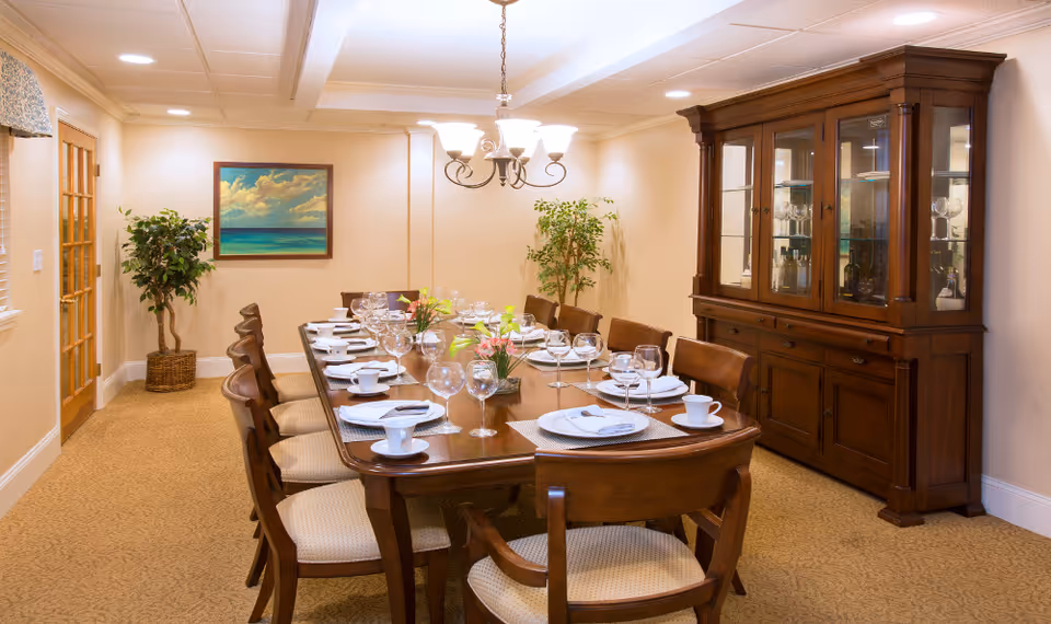 A formal dining room with a long wooden table set for twelve people. The table is arranged with white plates, cups, napkins, and wine glasses. There are two small flower arrangements in the center of the table. The room has beige walls, a carpeted floor, a wooden china cabinet filled with glassware, two potted plants, a framed painting of a seascape on the wall, and a chandelier hanging above the table.