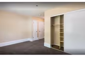 Empty room with beige walls and dark carpet flooring featuring an open closet with built-in shelves and hanging space, two closed white doors, and white baseboards.