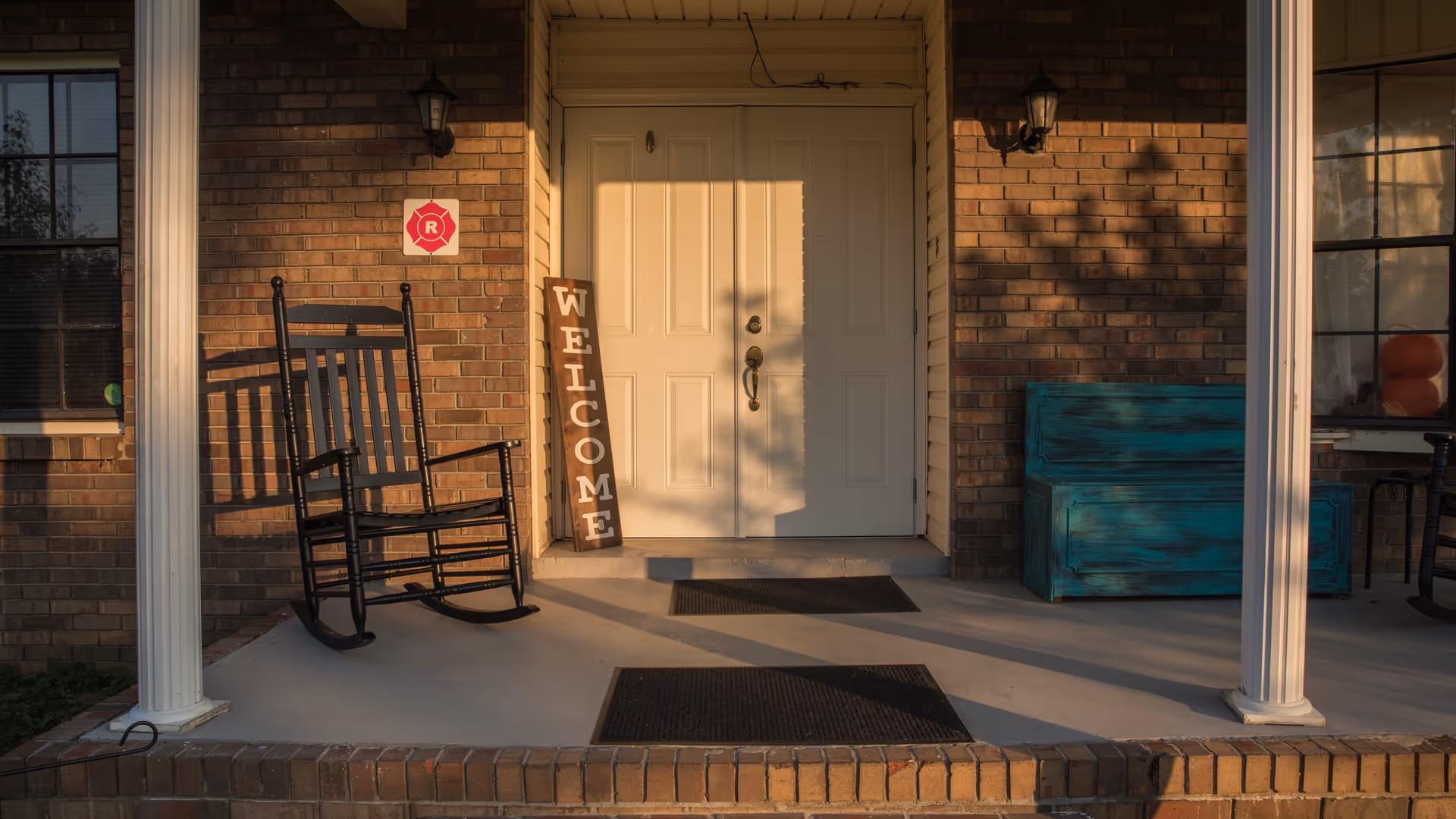 Front porch of a brick building with a white double door entrance. There is a black rocking chair on the left side, a vertical wooden sign that says 'WELCOME' leaning against the wall, and a teal wooden bench on the right side. Two white columns frame the porch, and there are two black wall-mounted lanterns on either side of the door.