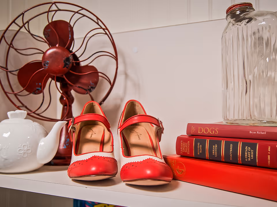 Pair of red-and-white women's shoes displayed on a shelf alongside a vintage red fan, teapot, stacked books and a glass jar.