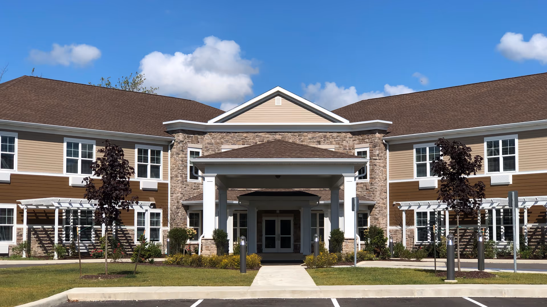 Front exterior view of a two-story senior living facility building with beige and brown siding, stone accents, a covered entrance supported by white columns, small trees, and a well-maintained lawn under a blue sky with some clouds.