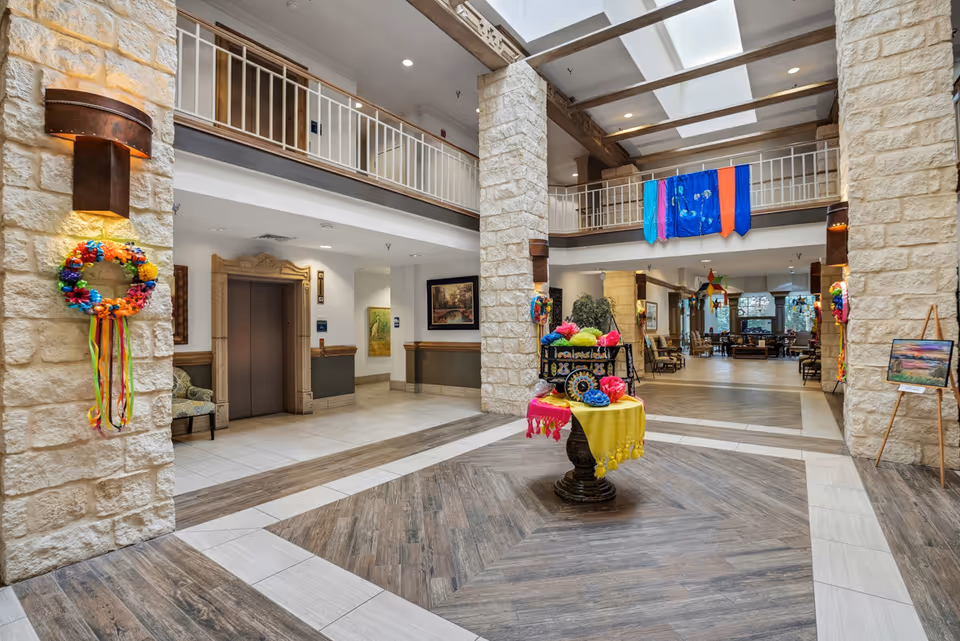 Spacious and well-lit interior lobby area with stone pillars, a decorative table with colorful fabric and flowers in the center, and an elevator on the left side. The space features a high ceiling with skylights, a second-floor balcony railing, wall sconces, and various pieces of artwork and seating areas in the background.