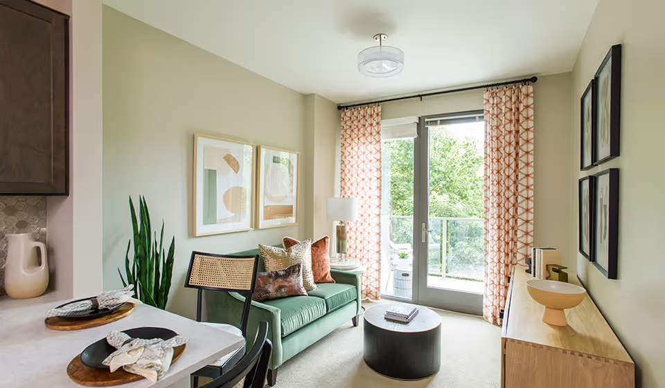 A cozy living room area with a green sofa adorned with multiple decorative pillows, a round black coffee table with books on it, and a wooden sideboard with decorative items. The room has large glass doors with patterned curtains that open to a balcony with greenery outside. There is a dining counter with place settings and chairs visible on the left side.