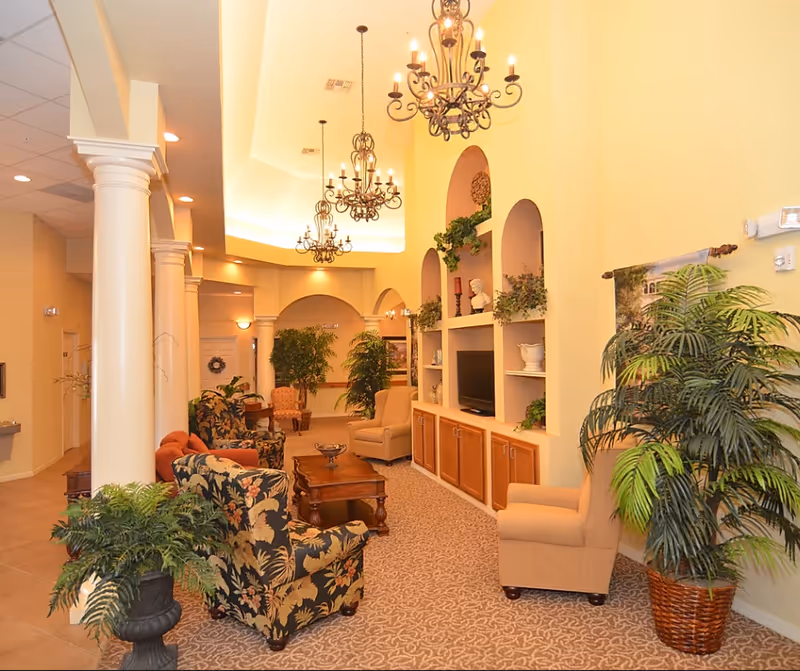 A spacious and warmly lit living room area in a senior living facility featuring multiple upholstered armchairs with floral and solid patterns, a wooden coffee table, built-in shelves with decorative items and a television, several large potted plants, and ornate chandeliers hanging from a high ceiling.