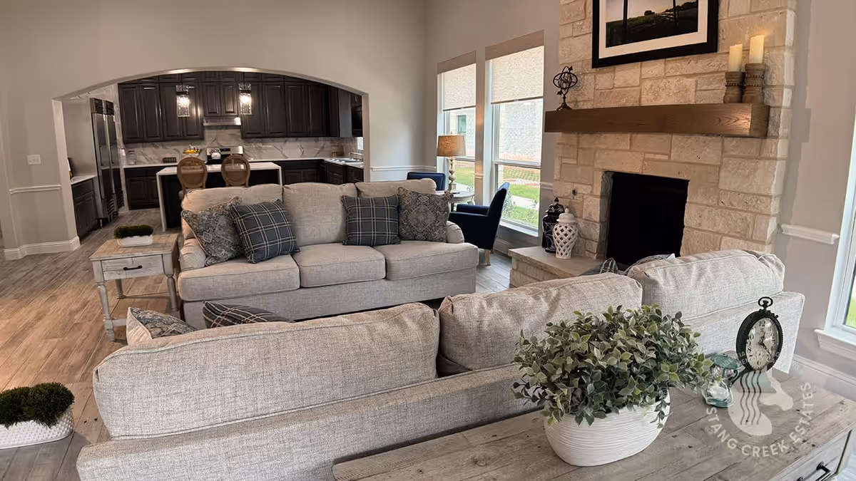 A cozy living room with two beige sofas adorned with patterned cushions, a wooden side table with a potted plant and a clock, a stone fireplace with candles and decorative items on the mantel, and large windows letting in natural light. The living room opens into a kitchen area with dark cabinets, a marble backsplash, and a kitchen island with two chairs.