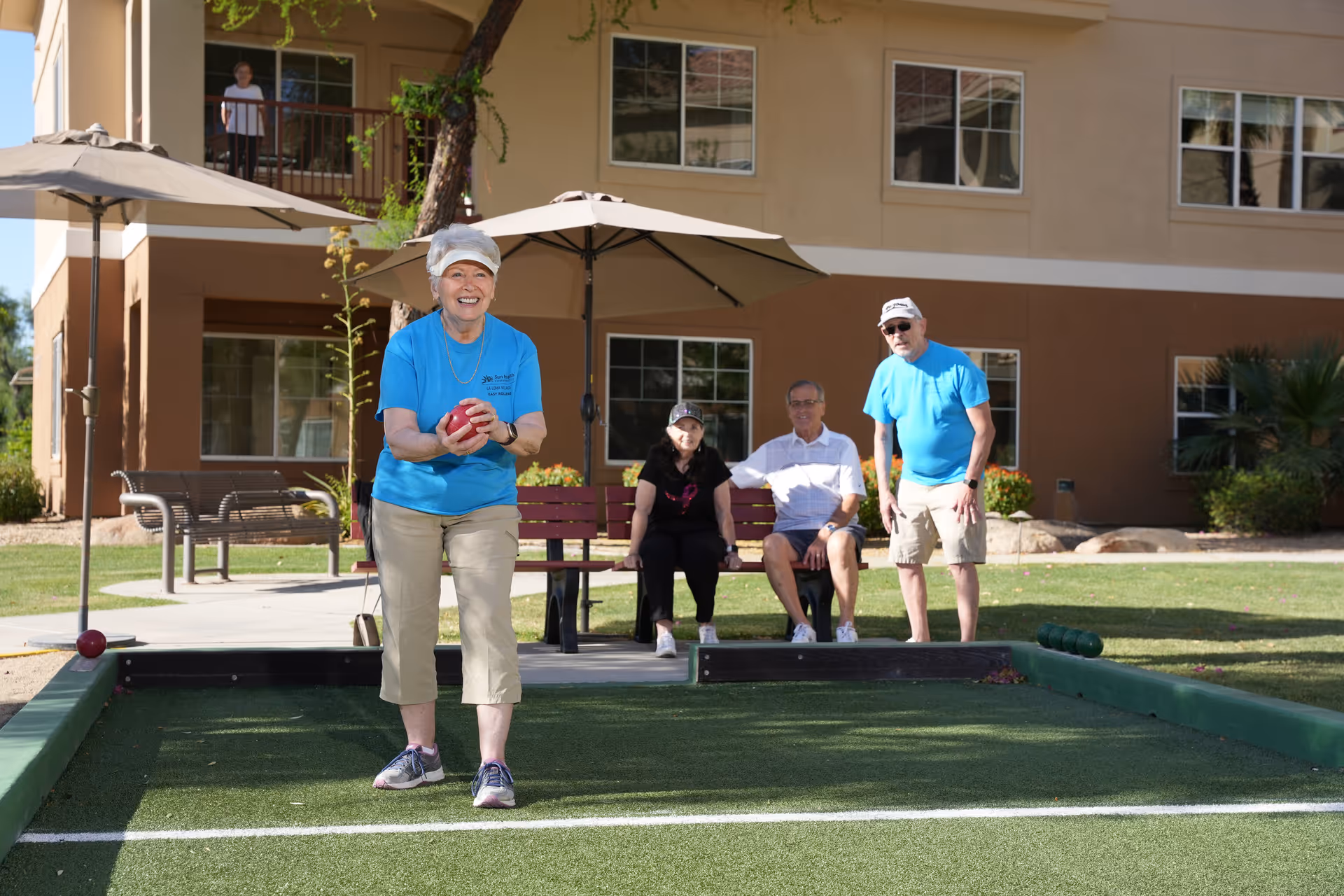 A group of elderly people enjoying a game of bocce ball on a green court outside a residential building. One woman in a blue shirt and beige pants is holding a red bocce ball and smiling, while two people sit on a bench and another man in a blue shirt and beige shorts stands nearby. The building has multiple windows and balconies, with umbrellas providing shade over benches.