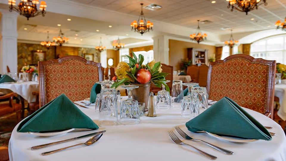 A dining table set for a meal in an elegant dining room with white tablecloth, green folded napkins, glassware, silverware, and a centerpiece with flowers and fruit. The background shows more tables and chairs with warm lighting from chandeliers.