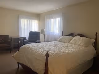 A bedroom with a large bed covered in a white quilt, two pillows, a wooden headboard and footboard. The room has two windows with sheer white curtains, a floor lamp between the windows, and two chairs placed against the walls.