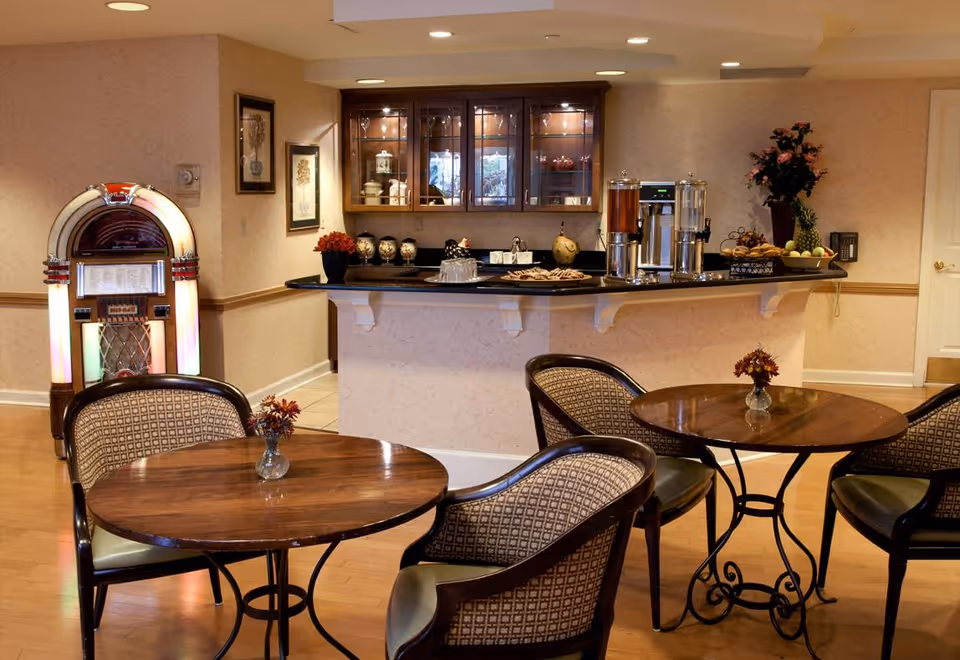 Cozy dining/lounge area with round wooden tables and upholstered chairs, a countertop buffet with drink dispensers, and a jukebox.