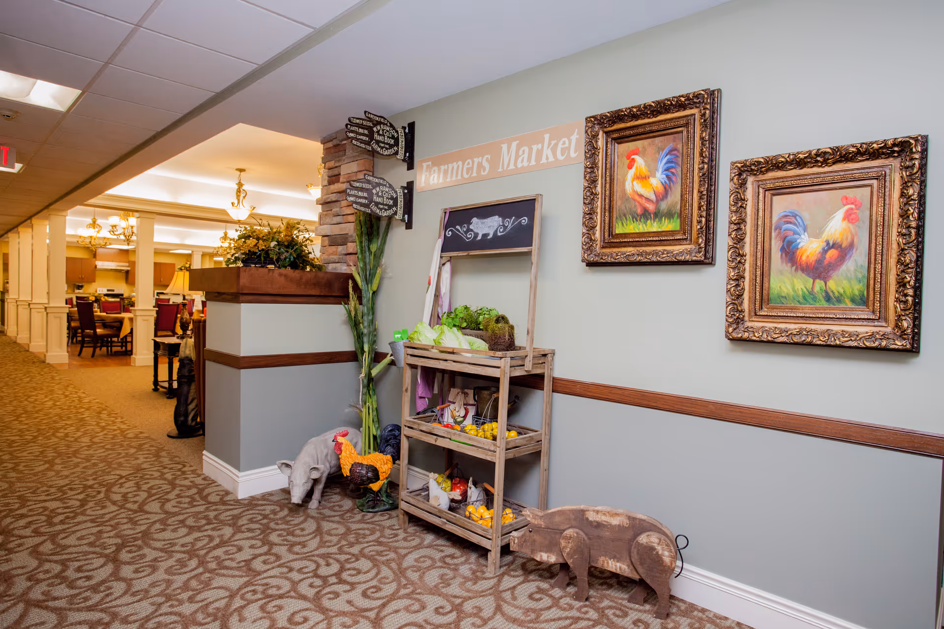 Interior hallway of a senior living facility decorated with a small wooden Farmers Market display holding artificial vegetables and fruits. The wall behind the display has two framed paintings of roosters and a sign that reads 'Farmers Market'. The hallway leads to a dining area with tables and chairs visible in the background.