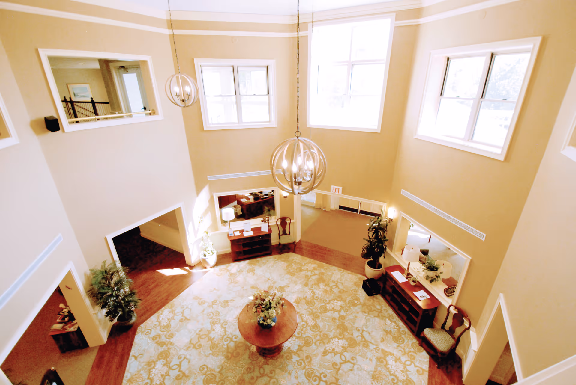 Bright two-story lobby with hanging spherical chandeliers, a central round table with flowers, seating, potted plants, and multiple windows.