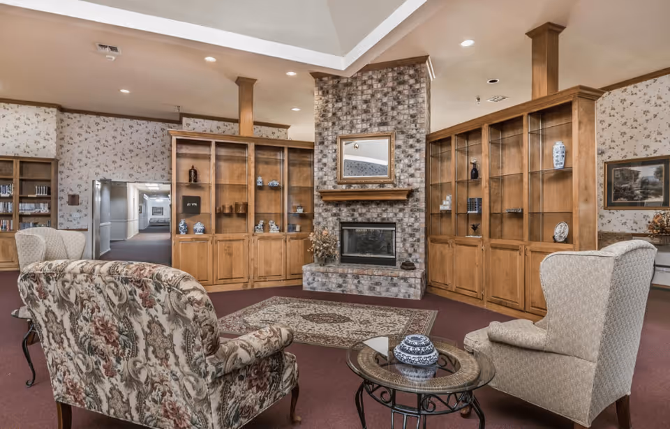 Cozy common room with patterned armchairs, a glass-top coffee table, and a brick fireplace flanked by wooden bookcases.