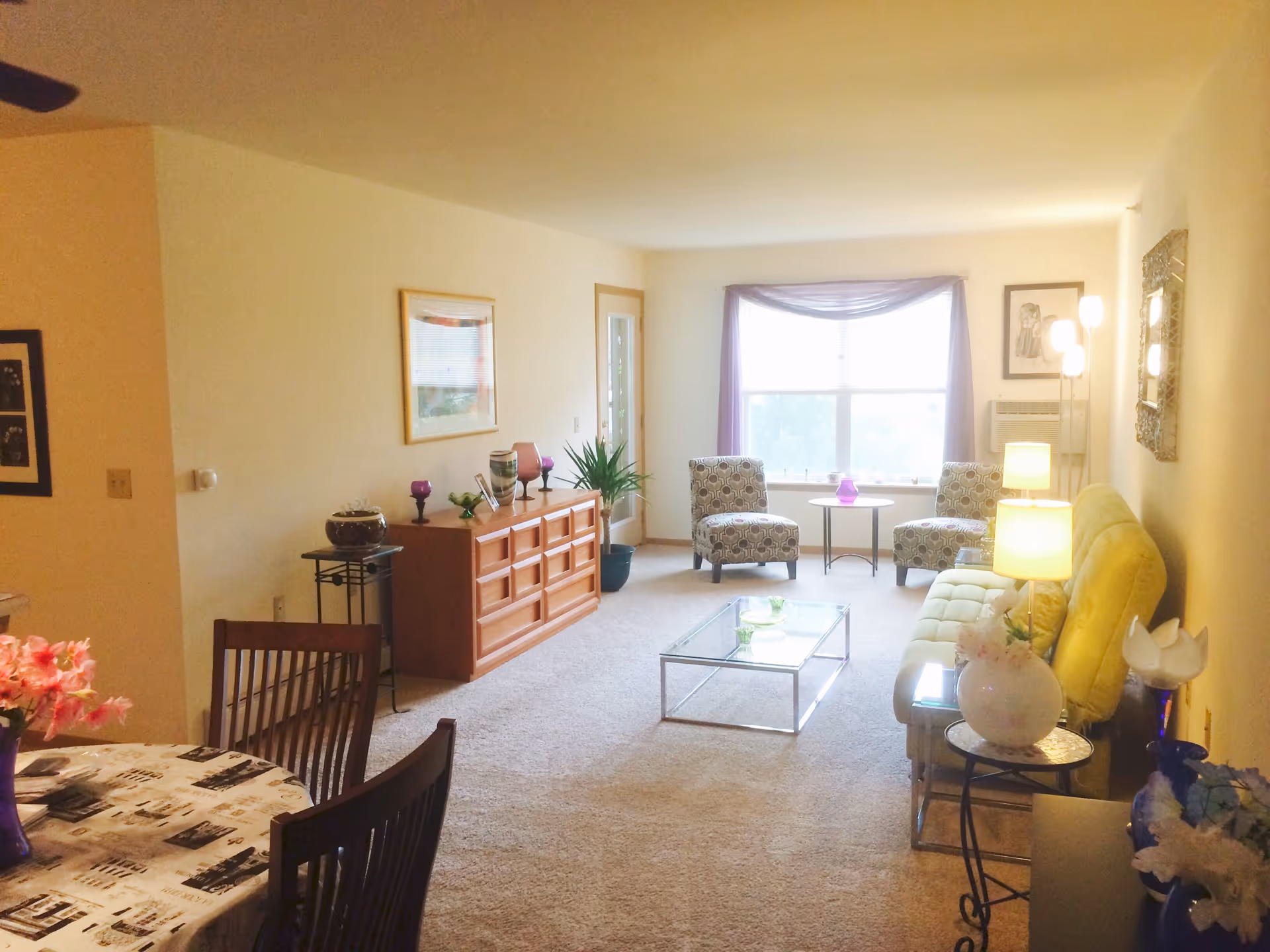 A bright living room with beige walls and carpet. The room features a yellow sofa with a lamp on a side table, two patterned armchairs near a large window with purple curtains, a glass coffee table, and a wooden dresser with decorative items. A dining table with chairs and a floral centerpiece is partially visible in the foreground.