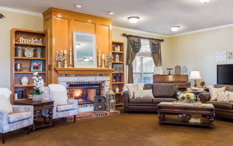 A cozy living room with a lit brick fireplace surrounded by wooden shelves filled with decorative items and books. There are two upholstered armchairs with a small wooden side table between them on the left, and two brown leather sofas with cushions on the right. A wooden coffee table with a floral centerpiece sits in front of the sofas. The room has beige walls, a window with brown curtains, and ceiling lights.