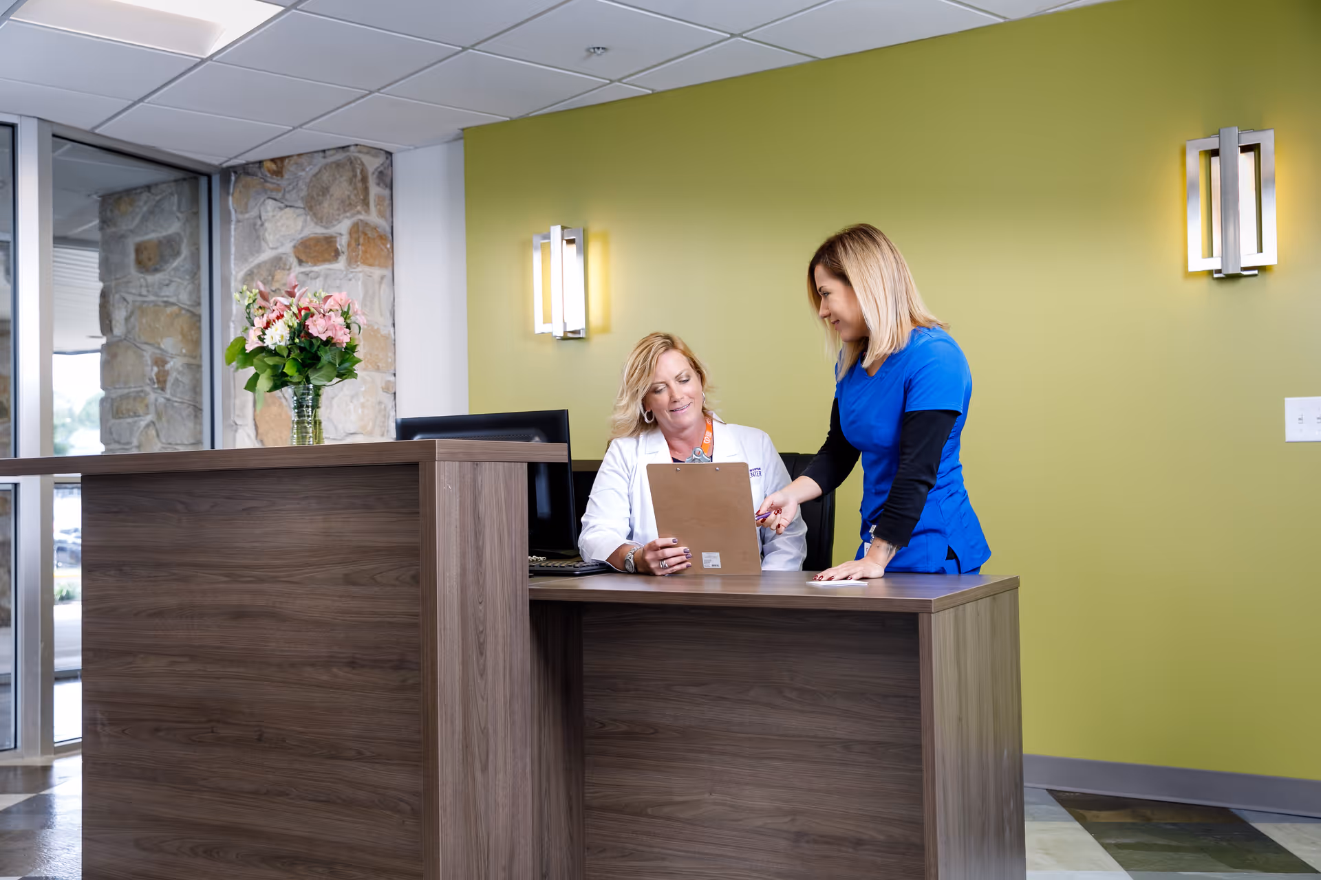 Two women at a reception desk in a facility. One woman in a white coat is seated and holding a clipboard, while the other woman in blue scrubs is standing and pointing at the clipboard. The background features a green wall with modern wall sconces and a vase of flowers on the desk.