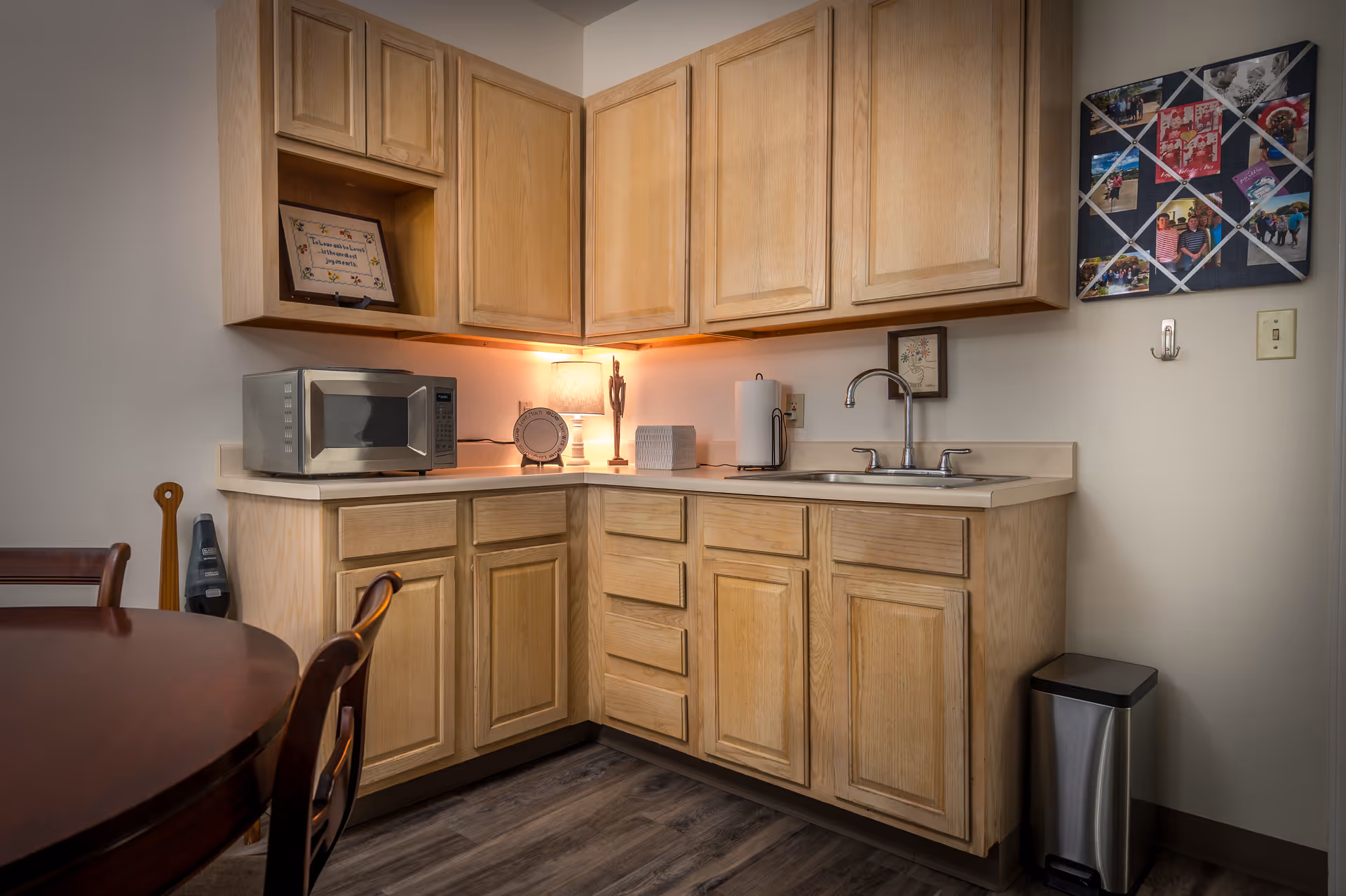 A small kitchen area with light wood cabinets, a countertop with a microwave, a lamp, a paper towel holder, and a sink. There is a round wooden table with chairs partially visible on the left side. On the wall to the right, there is a photo collage and a small framed picture. A stainless steel trash can is placed on the floor near the wall.