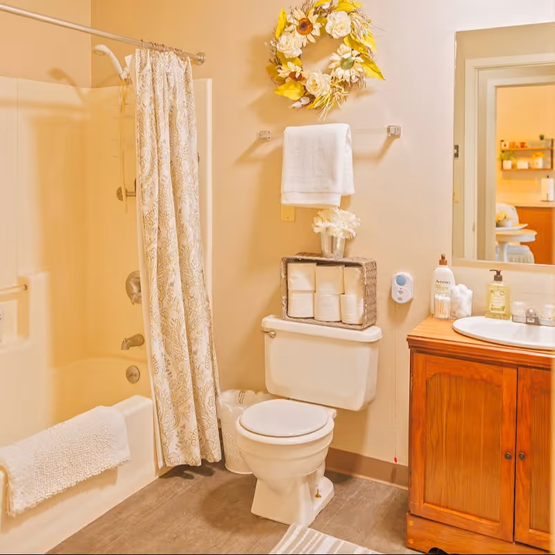 Well-lit bathroom featuring a shower/tub with a patterned curtain, a toilet topped with rolled toilet paper, a wooden vanity sink, and a decorative wreath on the wall.