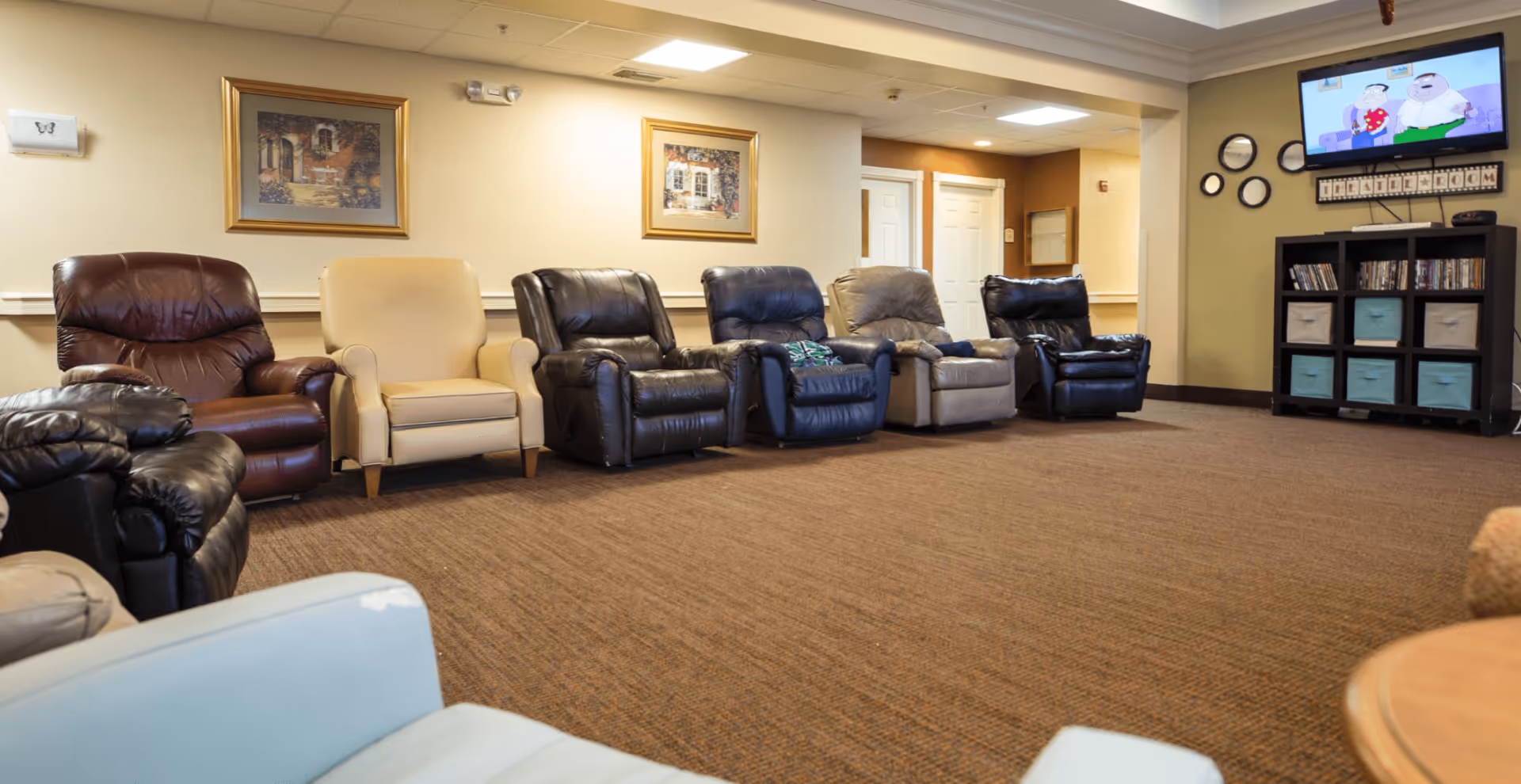 A spacious common room with a row of recliner chairs facing a wall-mounted TV and shelving with storage bins.