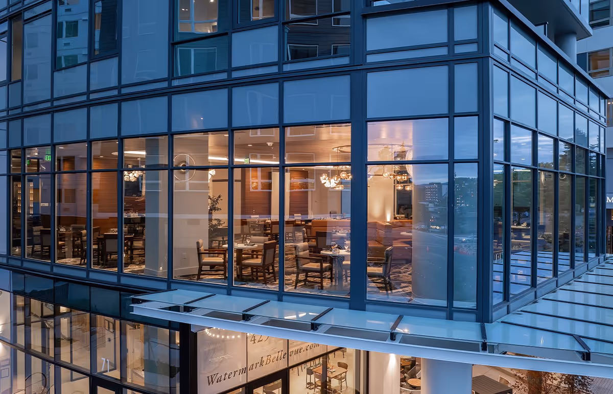 Exterior view of a modern building with large glass windows revealing a well-lit dining area inside, featuring tables and chairs arranged neatly, with chandeliers hanging from the ceiling.