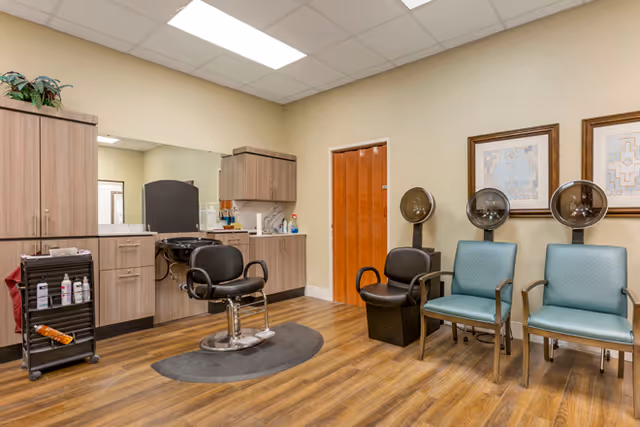 Interior of a salon room in a senior living facility with a black salon chair in front of a counter and mirror, two hair drying stations with black chairs, and two blue chairs against the wall. The room has wooden flooring, beige walls, and framed artwork on the wall.